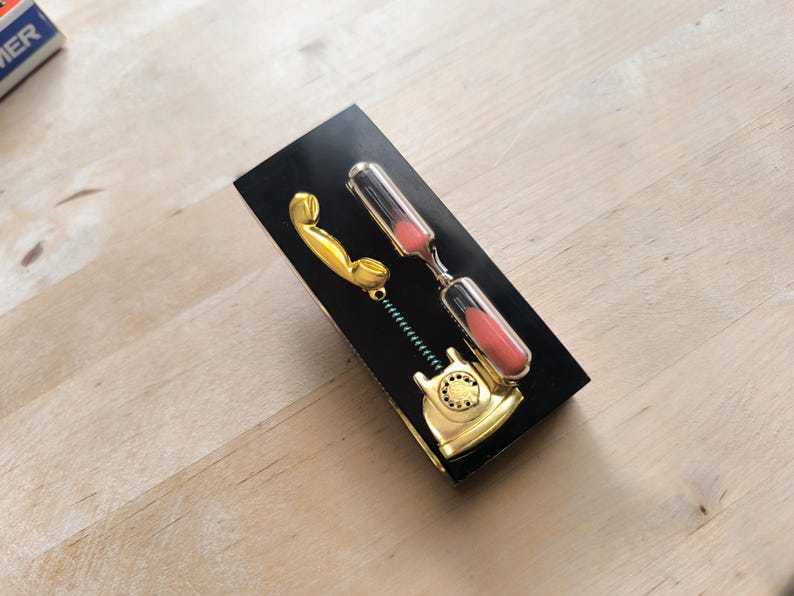 May include: A gold-colored phone receiver with a black hourglass attached to it. The hourglass is filled with pink sand. The phone receiver and hourglass are on a black background.