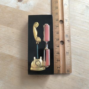 May include: A gold-colored rotary phone with a black background and a pink hourglass. The phone is attached to a spring.
