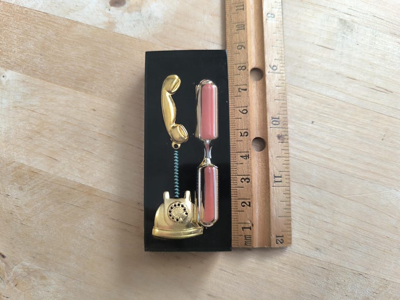 May include: A gold-colored rotary phone with a black background and a pink hourglass. The phone is attached to a spring.
