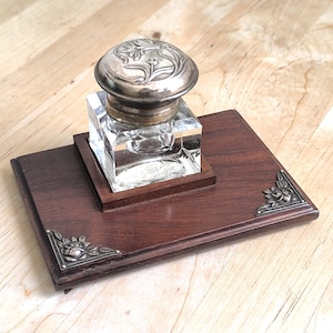 May include: An antique inkwell set featuring a clear glass inkwell with a silver-colored lid, resting on a dark wooden base. The base has decorative metal corner accents. A classic writing accessory.
