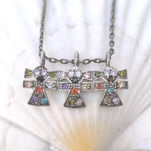 Peut inclure: Un collier en argent avec trois pendentifs en forme de personnages ornés de pierres précieuses colorées. Chaque personnage a une tête avec des pierres claires et un corps avec des gemmes de couleurs variées. Le collier est présenté sur une surface blanche texturée.