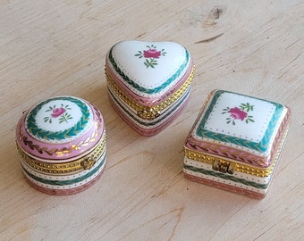 May include: Three decorative porcelain trinket boxes in different shapes: round, heart, and square. Each box features a floral design on the lid, with pink, green, and gold accents. The boxes have a hinged closure and are set against a light wood surface.