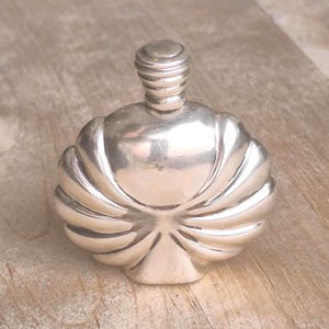 Puede incluir: Frasco de perfume plateado con diseño en forma de concha. El frasco tiene una base redondeada y un tapón cilíndrico estriado. El frasco es de metal y está sobre una superficie de madera.