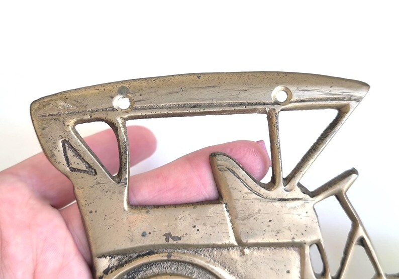May include: Vintage brass train-shaped wall decor. The decorative piece features a detailed design of a train engine with cut-out windows and a circular front. The brass has a weathered appearance, suggesting age and use.