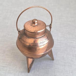 Puede incluir: Una pequeña olla de metal color cobre con tapa y asa. La olla tiene tres patas y una banda decorativa grabada alrededor del centro. El asa es arqueada y la tapa tiene un pequeño pomo.
