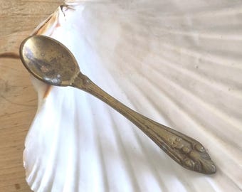 May include: A small, antique brass spoon with a rounded bowl and a decorative handle. The spoon is tarnished, showing signs of age and use. The handle features an embossed floral design. The spoon is resting on a white, textured surface.
