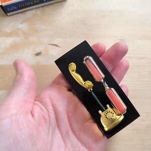 May include: A gold-colored rotary phone with a pink hourglass attached to the receiver. The phone and hourglass are mounted on a black background.