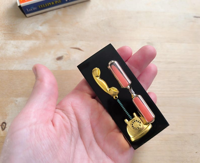 May include: A gold-colored rotary phone with a pink hourglass attached to the receiver. The phone and hourglass are mounted on a black background.