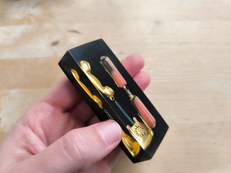 May include: A black resin paperweight with a gold rotary phone and an hourglass with pink sand. The phone and hourglass are embedded in the resin.