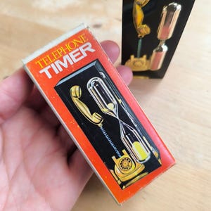 May include: A red box with the words "Telephone Timer" on it. The box shows a black rotary phone with a gold receiver and an hourglass. The hourglass is filled with yellow sand.