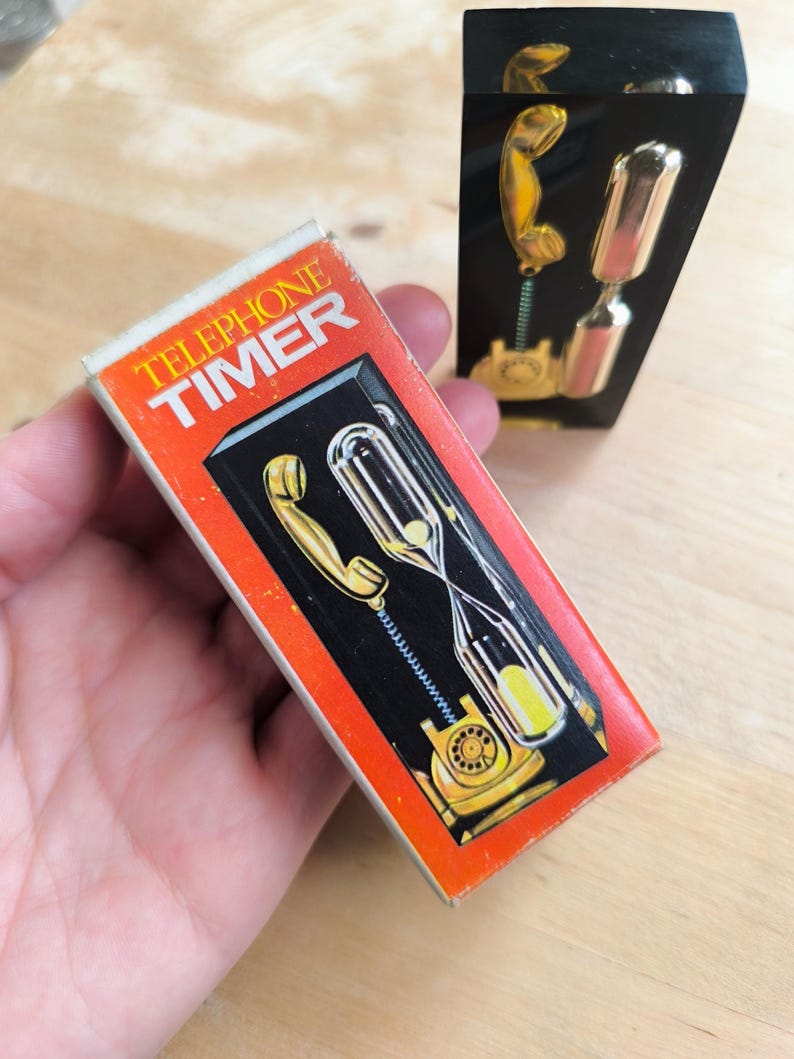 May include: A red box with the words "Telephone Timer" on it. The box shows a black rotary phone with a gold receiver and an hourglass. The hourglass is filled with yellow sand.