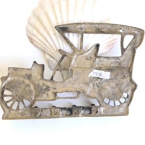 May include: A vintage-style metal car wall hanging with a weathered, antique finish. The car is a classic open-top design with spoked wheels and a detailed body. A seashell is in the background. The tag on the car reads "17a".