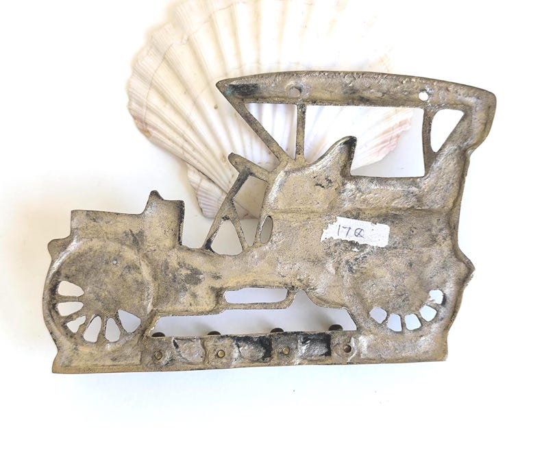 May include: A vintage-style metal car wall hanging with a weathered, antique finish. The car is a classic open-top design with spoked wheels and a detailed body. A seashell is in the background. The tag on the car reads "17a".