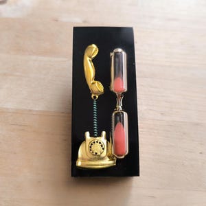 May include: A gold-colored phone with a black hourglass on a black background. The hourglass is filled with pink sand.