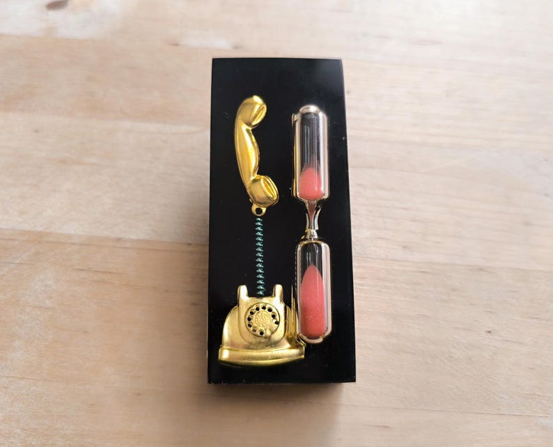 May include: A gold-colored phone with a black hourglass on a black background. The hourglass is filled with pink sand.