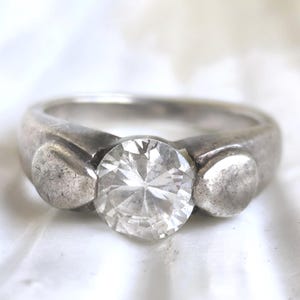 May include: A silver ring with a large, clear, round gemstone as the centerpiece. The ring has a thick band with two small, round accents on either side of the gemstone. The ring is set against a white background.