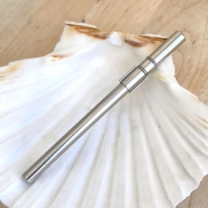 Puede incluir: Una pajita de acero inoxidable con un diseño segmentado. La pajita es cilíndrica y tiene un acabado metálico pulido. La pajita está sobre una concha blanca. El fondo es una superficie de madera de color marrón claro.