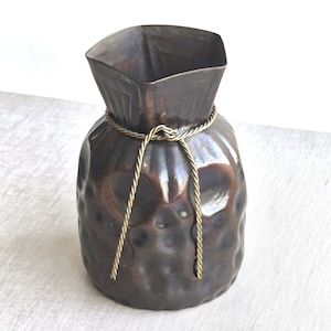 May include: A dark brown metal vase with a textured body and a square top. A gold-colored rope is tied around the neck of the vase. The vase has a unique, handcrafted appearance, suitable for home decor.