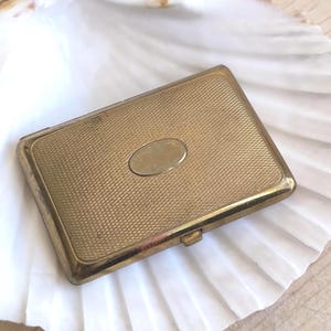 Può includere: Un astuccio rettangolare vintage in metallo dorato con un motivo strutturato a tratteggio incrociato. Un accento ovale è centrato sul coperchio. L'astuccio è appoggiato su una superficie bianca strutturata, forse una conchiglia.