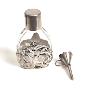 Puede incluir: Un frasco de perfume de vidrio transparente con una base floral plateada y una tapa plateada. Un embudo plateado separado está al lado del frasco. El frasco tiene forma ovalada.