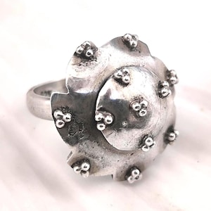 Op de afbeelding: Een zilveren ring met een uniek, getextureerd ontwerp. De ring heeft een grote, gestileerde bloemvorm met een spiraalvormig midden en kleine, verhoogde, parelachtige details. De band is glad en contrasteert met de sierlijke bovenkant.