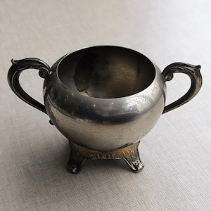 Puede incluir: Una jarra de crema de plata con cuerpo redondeado y dos asas. La jarra tiene una base decorativa con tres patas.