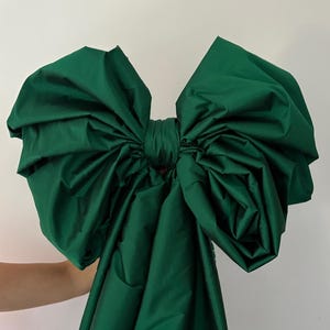 May include: A large, emerald green bow made of a shiny fabric. The bow is voluminous, with multiple folds and layers, and is tied in the center. The fabric has a slight sheen, reflecting light. The bow is against a plain white background.