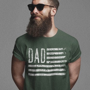 May include: A green t-shirt with a white distressed American flag design and the word "DAD" printed on it.