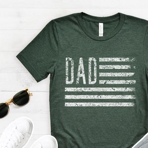 May include: A dark green t-shirt with a white distressed graphic of an American flag with the word "DAD" in place of the stars.