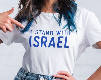 May include: A woman wearing a white t-shirt with the text "I stand with Israel" in blue lettering. She is pointing at the shirt with her right hand.