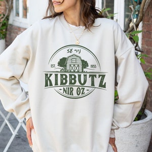 May include: A beige sweatshirt with a green graphic design of a barn with the text "Kibbutz Nir Oz" and "Est. 1955".