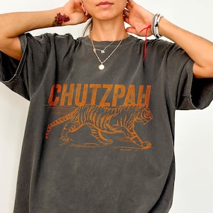 May include: A dark gray oversized t-shirt with an orange graphic design. The design features the word 'CHUTZPAH' in bold letters above an orange tiger illustration. The shirt has short sleeves and a relaxed fit, suggesting a casual style.
