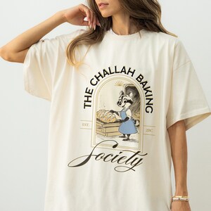 K&ouml;nnte beinhalten: Cremefarbenes T-Shirt mit einem Waschb&auml;r-B&auml;cker-Motiv. Der Waschb&auml;r tr&auml;gt eine blaue Sch&uuml;rze und h&auml;lt Brot. Der Text "THE CHALLAH BAKING Society" und "EST. 2010" sind ebenfalls auf dem Shirt abgebildet. Das Shirt ist oversized.
