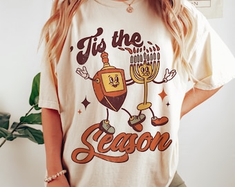 May include: A beige t-shirt with a vintage-style graphic of a dreidel and a menorah. The graphic says "'Tis the Season".