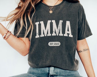 May include: A dark grey t-shirt with the text "IMMA EST. 2023" in white. The shirt has a distressed look and is being worn by a person with long brown hair.