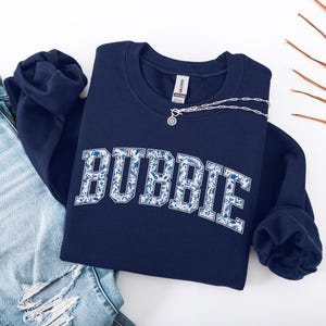 May include: Navy blue sweatshirt with the word "BUBBIE" in a white and blue floral pattern. A silver necklace with a small pendant rests on the sweatshirt. Light blue denim jeans are visible on the left.