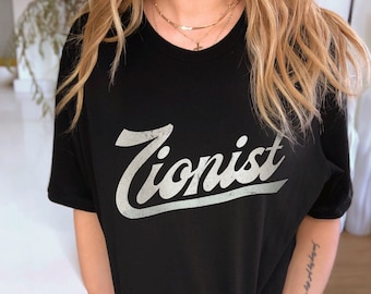 Op de afbeelding: Zwart T-shirt met het woord "Zionist" in een verweerde, witte retro-lettertype. Het shirt heeft een ronde hals en lijkt gemaakt van een zacht materiaal. Het model draagt gouden kettingen.