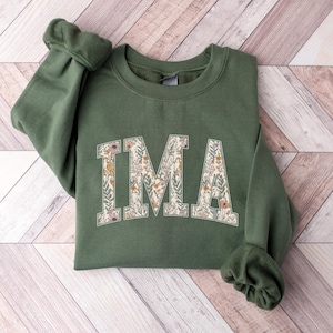 Könnte beinhalten: Ein olivgrünes Sweatshirt mit den Buchstaben "IMA" in einem floralen Design. Das Sweatshirt liegt gefaltet auf einer hellbraunen und weißen Holzoberfläche. Die Ärmel sind umgeschlagen.