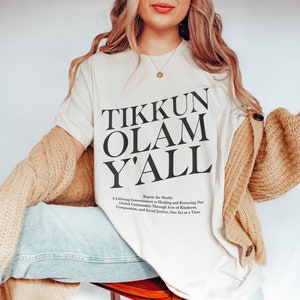May include: A white t-shirt with the text "Tikkun Olam Y'all" printed in black. The text is centred on the shirt and is surrounded by a smaller text that reads "Repair the World: A Lifelong Commitment to Healing and Restoring Our Global Community Through Acts of Kindness, Compassion, and Social Justice, One Act at a Time."