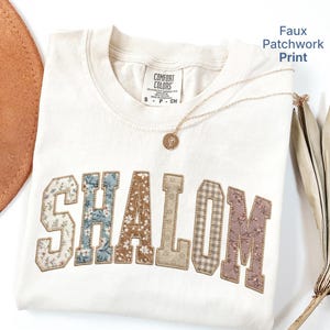 May include: Cream-colored Comfort Colors t-shirt with the word "SHALOM" in a faux patchwork print. The letters feature floral and plaid patterns in shades of blue, brown, and pink. A gold necklace and a brown hat are also visible.
