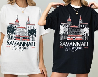 May include: White and black t-shirts featuring a graphic of Savannah, Georgia. The design includes buildings, a trolley, and the text "SAVANNAH Georgia ESTD. 1733". The shirts are oversized and have short sleeves.