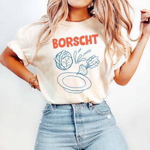 Peut inclure: T-shirt crème avec le mot "BORSCHT" en rouge, au-dessus d'un dessin bleu et blanc d'un bol et de légumes. Le t-shirt est porté avec un jean bleu clair.