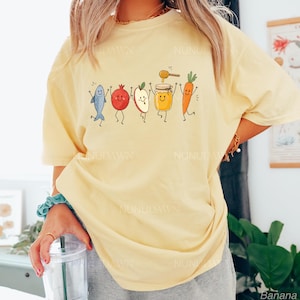 May include: A pale yellow short-sleeve t-shirt featuring a graphic print of a blue fish, a red pomegranate, a sliced apple, a jar of honey with a dipper, and an orange carrot, all depicted with smiling faces and arms raised in a line.