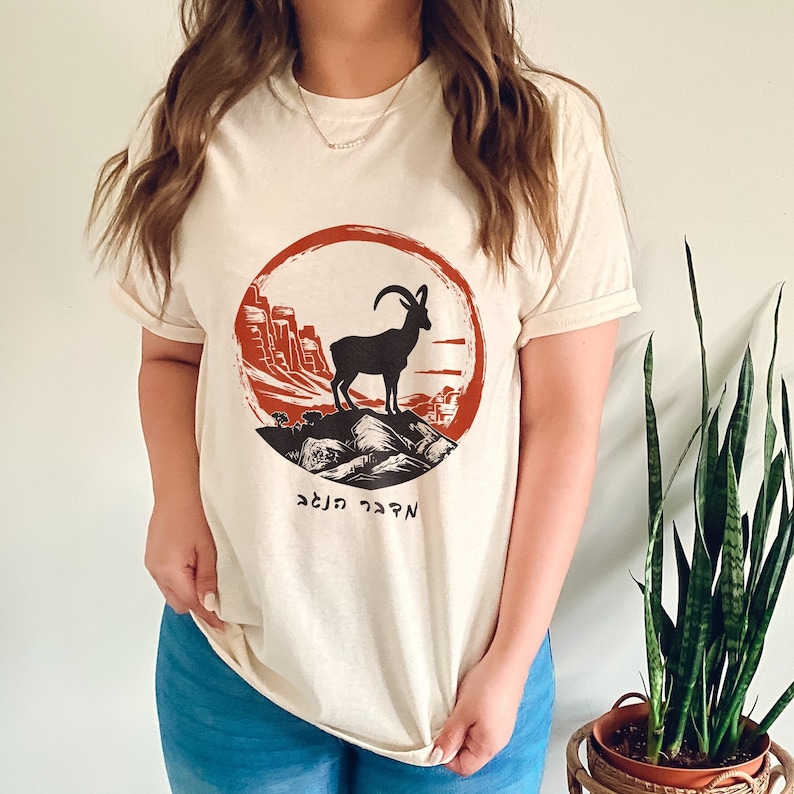 May include: Cream-colored t-shirt featuring a circular graphic with a black mountain goat silhouette atop a mountain range, set against a red and orange backdrop. The shirt includes Hebrew text below the graphic. The shirt is made of a soft material.