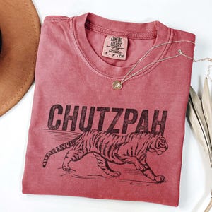 May include: A red-toned t-shirt with the word "CHUTZPAH" printed in black, along with a tiger illustration. The shirt has a crew neck and a small tag at the neckline. A gold necklace with a pendant is draped across the shirt.