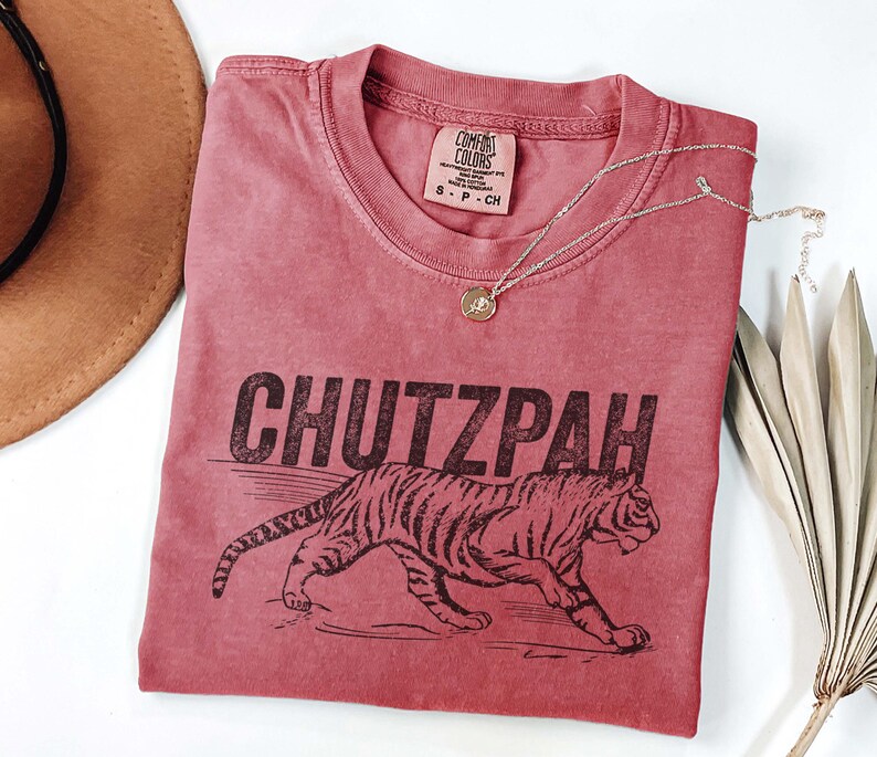 May include: A red-toned t-shirt with the word "CHUTZPAH" printed in black, along with a tiger illustration. The shirt has a crew neck and a small tag at the neckline. A gold necklace with a pendant is draped across the shirt.