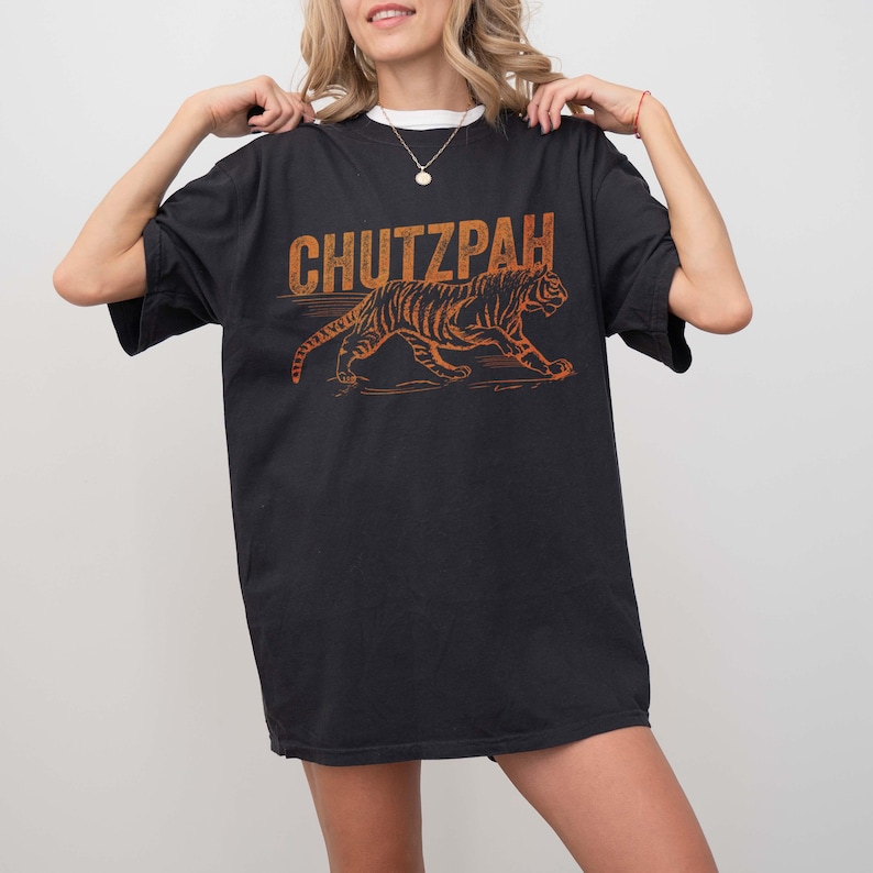 May include: Black t-shirt with the word "CHUTZPAH" in orange block letters above an orange tiger graphic. The tiger is walking to the right. The shirt is being held up by a person.