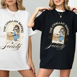 K&ouml;nnte beinhalten: Wei&szlig;e und schwarze T-Shirts mit dem Text "THE CHALLAH BAKING Society" und einer Illustration eines B&auml;ckerracoon. Das Design befindet sich in einem goldenen Rahmen mit den Worten "EST. 2017". Die Shirts sind kurz&auml;rmelig und haben eine lockere Passform.