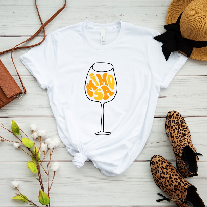 May include: White t-shirt with a graphic of a wine glass with the word "MIMOSA" inside the glass in a retro font.