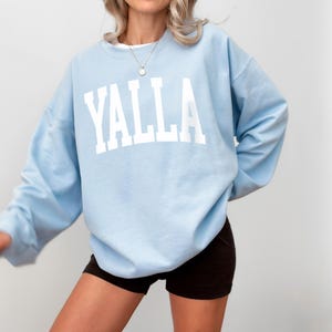 May include: A light blue sweatshirt with the word "YALLA" printed in white on the front. The sweatshirt is oversized and has a crew neck.